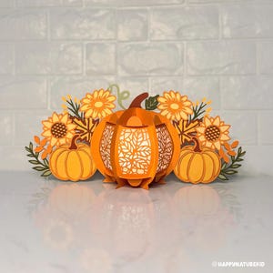 May include: An orange paper lantern with a leaf design, flanked by two smaller pumpkins and sunflowers. The lantern is the focal point, with intricate cutouts. The arrangement evokes a fall or harvest theme.