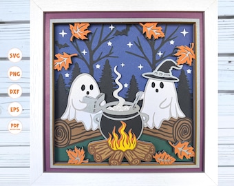 Op de afbeelding: Een ingelijst Halloween-thema papierkunstwerk. Twee witte geesten verzamelen zich rond een ketel, met een kampvuur eronder. De achtergrond is een donkerblauwe nachtscène met bomen, sterren en oranje bladeren. De lijst is wit.