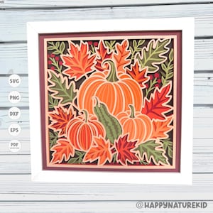 May include: A square framed wall art piece featuring a layered paper cut design of three pumpkins and a gourd surrounded by autumn leaves in shades of orange, red, and green. The artwork is set within a white frame and includes small red berries.