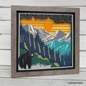 Puede incluir: Una obra de arte en papel enmarcada que representa una escena de monta&ntilde;a con un oso negro en primer plano. Las monta&ntilde;as est&aacute;n superpuestas en tonos de azul, verde y blanco, con un sol amarillo poni&eacute;ndose detr&aacute;s de ellas. El texto "Great Smoky Mountains National Park" est&aacute; en la parte superior de la imagen.