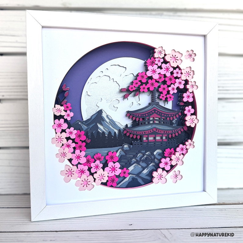 Puede incluir: Una obra de arte en papel enmarcada en blanco con un dise&ntilde;o en 3D en capas que representa una pagoda japonesa, flores de cerezo y una luna llena. La escena se desarrolla en un cielo morado y una cordillera.