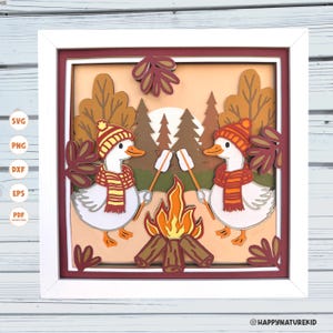 May include: A framed autumn-themed paper art piece. Two white geese wearing hats and scarves roast marshmallows over a campfire. The background features trees, a sun, and fall leaves. The frame is white.