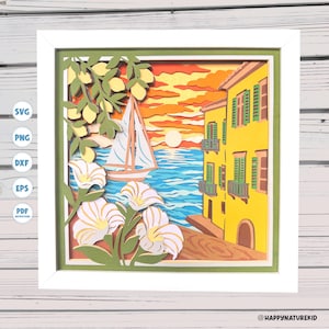 May include: A framed paper art piece showing a coastal scene. The artwork features a yellow building with green shutters, a sailboat on blue water, and white flowers. The background shows a sunset with orange and yellow hues.