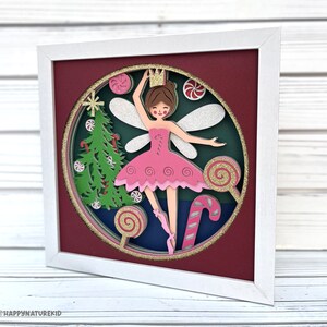 May include: A papercraft framed artwork featuring a ballerina fairy with pink wings and a pink tutu, surrounded by candy canes, lollipops, and a Christmas tree. The artwork is set against a red background and has a white frame.