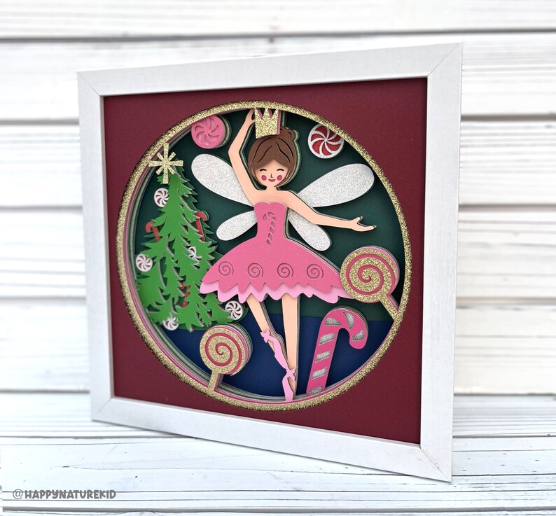 May include: A papercraft framed artwork featuring a ballerina fairy with pink wings and a pink tutu, surrounded by candy canes, lollipops, and a Christmas tree. The artwork is set against a red background and has a white frame.