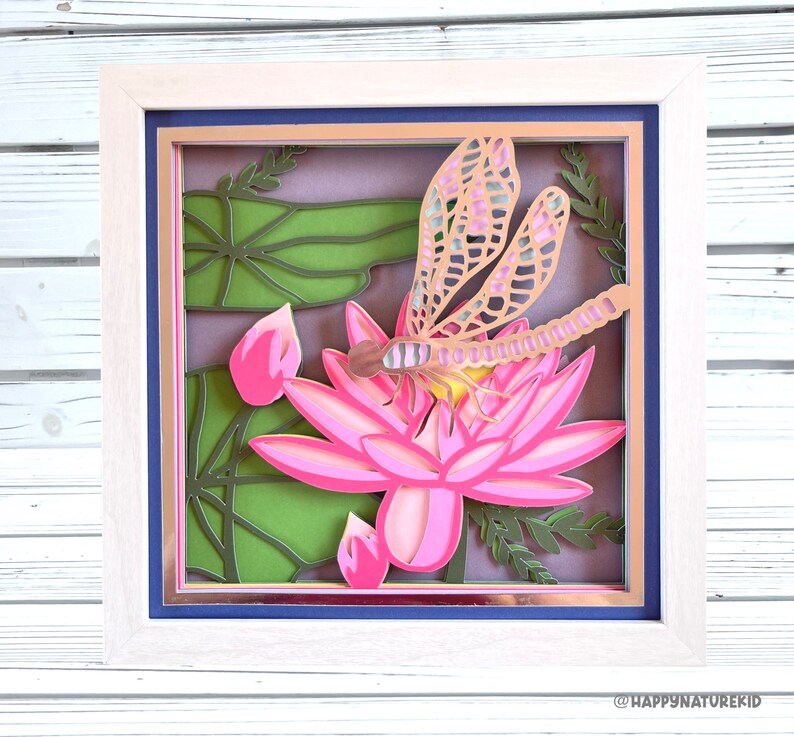 May include: A papercraft shadow box with a pink water lily and a dragonfly with gold accents. The background is a green and brown leafy design.