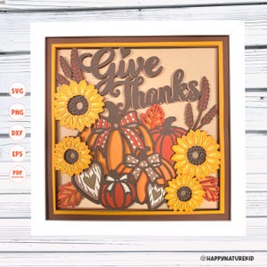 Puede incluir: Una pieza de arte en papel con capas en un marco blanco que presenta el texto Give Thanks en letra cursiva marrón oscuro. El diseño incluye girasoles amarillos, calabazas naranjas y rojas con lazos a cuadros y de lunares, y tallos de trigo.