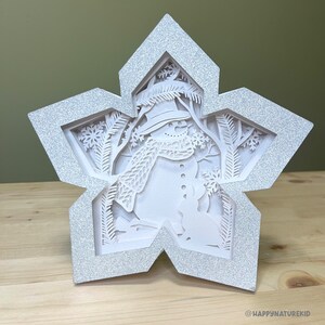 May include: A white paper snowflake ornament with a 3D cutout design of a snowman in a winter scene. The snowflake is decorated with glitter.