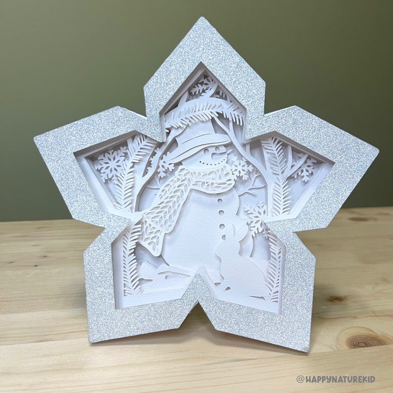 May include: A white paper snowflake ornament with a 3D cutout design of a snowman in a winter scene. The snowflake is decorated with glitter.