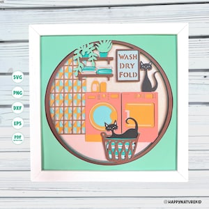 May include: A multi-layered paper craft art piece in a white frame featuring a laundry room scene with a washer, dryer, laundry basket, two black cats, and a sign that reads WASH DRY FOLD. The design includes shelves with towels and plants.