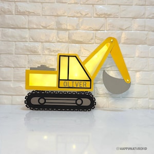 May include: A yellow and grey excavator-shaped wall light, with the name "OLIVER" displayed. The light features a black track base and a yellow arm with a grey scoop. It's set against a white brick wall.