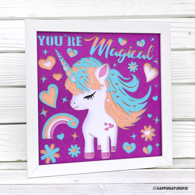 May include: White framed wall art with a purple glitter background and a white paper cut-out of a unicorn with a rainbow, stars, and hearts. The text "You're Magical" is at the top of the image.