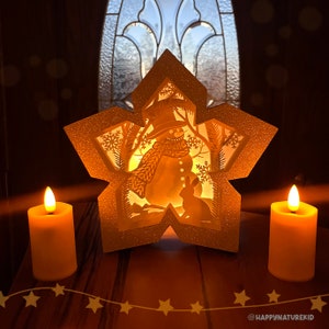 May include: A white star-shaped paper lantern with a cut-out design of a snowman and a rabbit. The lantern is illuminated from within, casting a warm glow. Two candles are placed on a wooden surface in front of the lantern.