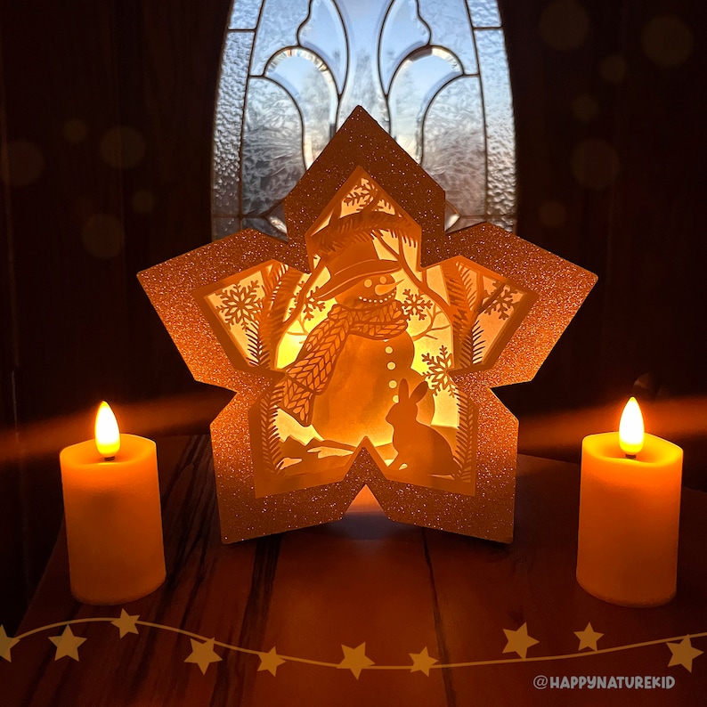 May include: A white star-shaped paper lantern with a cut-out design of a snowman and a rabbit. The lantern is illuminated from within, casting a warm glow. Two candles are placed on a wooden surface in front of the lantern.