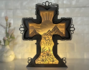 May include: A black wooden cross with a 3D design featuring a mountain scene and the words "He is Risen" in the center. The cross is illuminated from behind, creating a warm glow.
