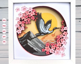 Puede incluir: Un marco blanco con un diseño redondo de papel recortado que presenta una grúa volando sobre una montaña con un sol amarillo y flores de cerezo rosas. El diseño está en capas y tiene un efecto 3D.