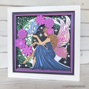 May include: A framed paper art piece featuring a fairy with brown hair and pink wings, seated on a crescent moon. The fairy is surrounded by pink flowers and a blue butterfly. The artwork is set against a black background with white stars.