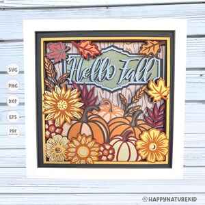 Op de afbeelding: Een ingelijst herfst-thema papierkunstwerk. Het kunstwerk toont gelaagde papieren uitsparingen van pompoenen, zonnebloemen en herfstbladeren in tinten oranje, geel en rood. De tekst "Hello Fall" is weergegeven.
