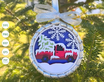 May include: A red truck with a Christmas tree in the back, covered in snow, is depicted on a round ornament with a silver glitter border. The ornament has a blue background with white snowflakes.