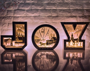 May include: A wooden JOY sign with a winter scene inside each letter. The J has a cafe and a toy store, the O has a ski lift and a horse-drawn carriage, and the Y has a church and a snowman.