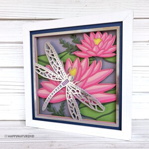 May include: A framed paper craft with a silver dragonfly and pink water lilies. The background is a layered paper design with green leaves and purple flowers.