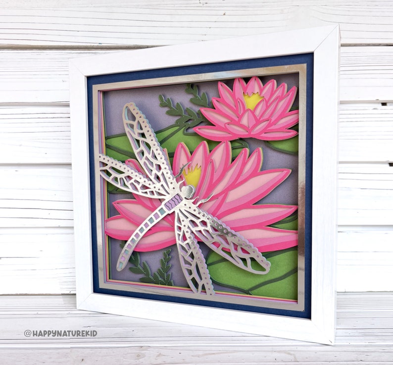 May include: A framed paper craft with a silver dragonfly and pink water lilies. The background is a layered paper design with green leaves and purple flowers.