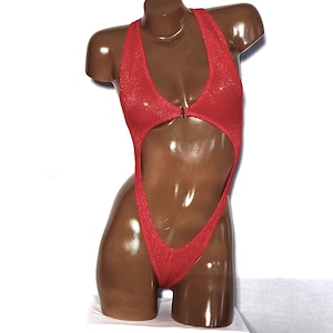 May include: A red, one-piece swimsuit with a halter neck and a high-cut design. The swimsuit features a keyhole cutout at the midriff and a small metal clasp. The fabric has a subtle shimmer, and the garment is displayed on a mannequin.