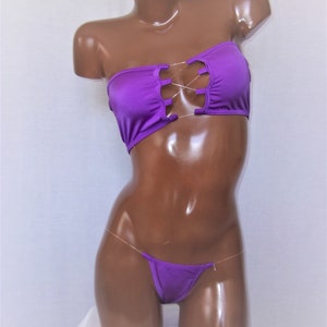 May include: Purple strapless bikini top with a criss-cross design and a matching purple bikini bottom with clear straps.