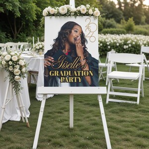 May include: A white easel sign with a floral arrangement at the top. The sign features a photo of a woman in a graduation gown with the text "Giselle's GRADUATION PARTY" and the year "2020". The sign is set up outdoors on a grassy lawn.