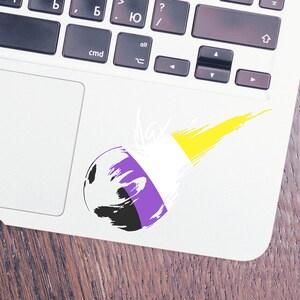 May include: A white laptop with a purple, white, and yellow sticker on the trackpad. The sticker is a stylized comet with the colors of the non-binary pride flag. The text "Max" is written in white above the comet.