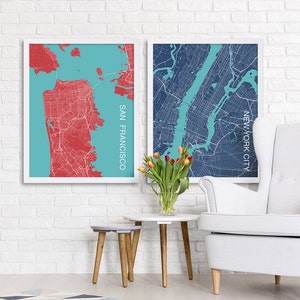 May include: Two framed city maps, one of San Francisco and one of New York City, are displayed on a white brick wall. The San Francisco map is in red and blue, while the New York City map is in blue and teal. A bouquet of pink tulips is in a clear vase on a gray and white side table with wooden legs. A white armchair with button tufting is in the foreground.