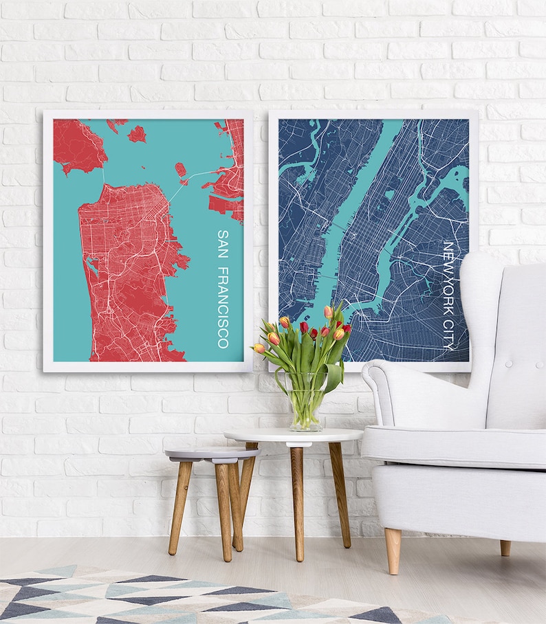 May include: Two framed city maps, one of San Francisco and one of New York City, are displayed on a white brick wall. The San Francisco map is in red and blue, while the New York City map is in blue and teal. A bouquet of pink tulips is in a clear vase on a gray and white side table with wooden legs. A white armchair with button tufting is in the foreground.