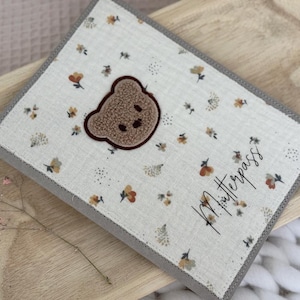 Könnte beinhalten: Ein hellfarbener Stoffbezug mit einem Teddybär-Applikation und floralen Mustern. Der Bezug hat einen grauen Rand und den Text "Mutterpass" in Schrift. Auf einem Holztablett platziert.