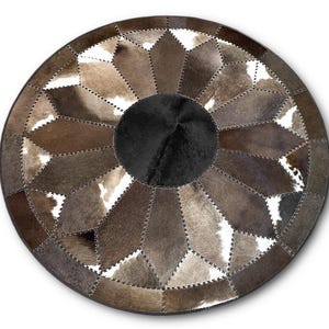 Op de afbeelding: Een ronde patchwork vloerkleed met een geometrisch ontwerp. Het kleed heeft verschillende tinten bruin, wit en zwart koeienhuid stukken die aan elkaar zijn gestikt. Het midden is een effen zwarte cirkel en de buitenrand is donkerbruin.