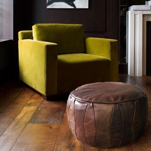 Könnte beinhalten: Ein brauner Leder-Pouf mit gestepptem Design steht vor einem grünen Samt-Sessel in einem Wohnzimmer.