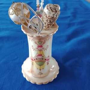 May include: A white ceramic vase with a floral design and a scalloped edge. The vase is filled with a variety of vintage Christmas ornaments, including a white ball with gold trim, a gold cone covered in sequins, and a clear glass ornament with a pink and white design.