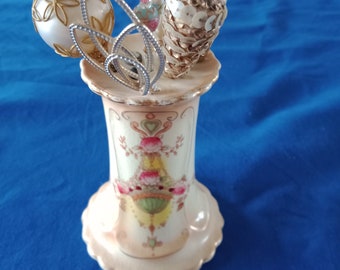 May include: A white ceramic vase with a floral design and a scalloped edge. The vase is filled with a variety of vintage Christmas ornaments, including a white ball with gold trim, a gold cone covered in sequins, and a clear glass ornament with a pink and white design.