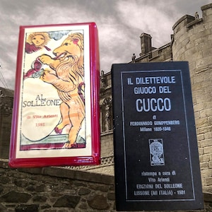 May include: Two rectangular card game boxes. The left box is red with a white label featuring a lion illustration, the sun, and the text AL SOLLEONE di Vito Arienti 1981. The right box is black with white text reading IL DILETTEVOLE GIUOCO DEL CUCCO.