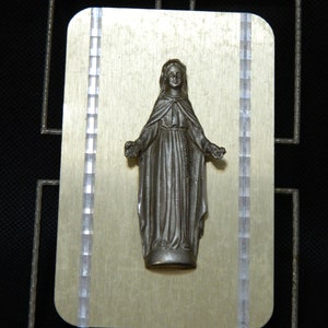 Peut inclure: Une statue argentée de la Vierge Marie en robe longue, debout sur une plaque dorée avec un cadre argenté.
