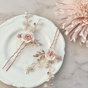 May include: Two rose gold hair pins with delicate pearl and crystal embellishments. Each pin features a pink rose with leaves and a wavy pin.