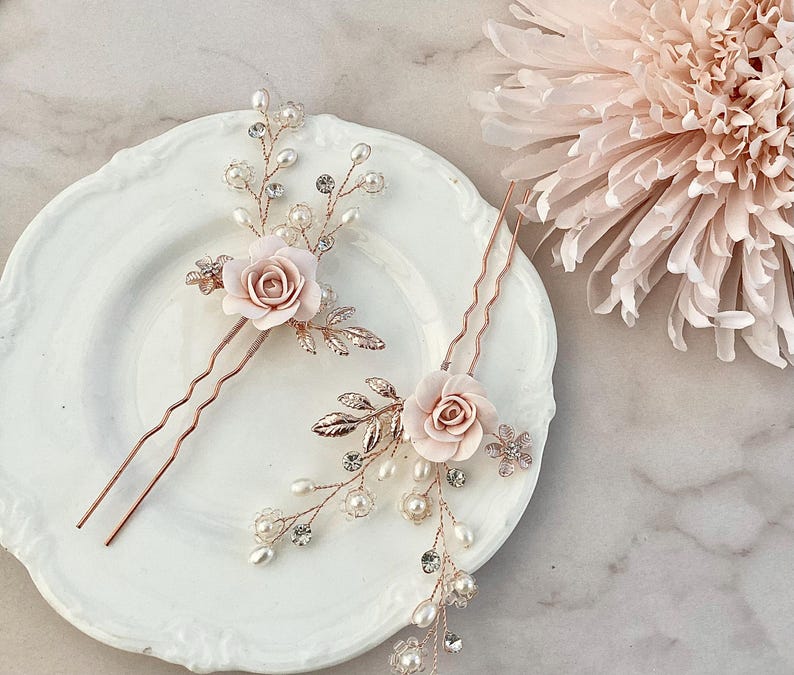 May include: Two rose gold hair pins with delicate pearl and crystal embellishments. Each pin features a pink rose with leaves and a wavy pin.