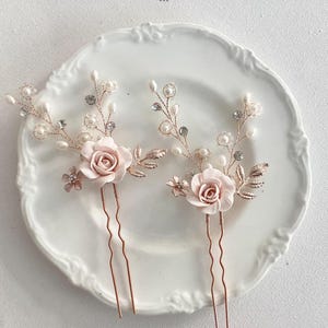May include: Two rose gold hair pins with delicate pearl and crystal accents. Each pin features a pink fabric rose and leafy details.
