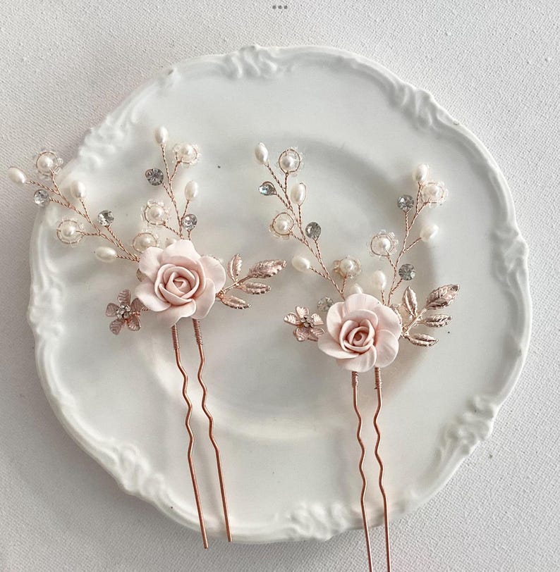 May include: Two rose gold hair pins with delicate pearl and crystal accents. Each pin features a pink fabric rose and leafy details.