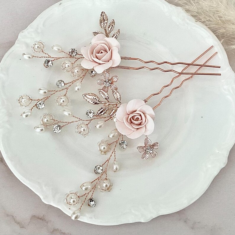 May include: Two rose gold hair pins with delicate pearl and crystal embellishments. Each pin features a pink fabric rose and a leafy design.