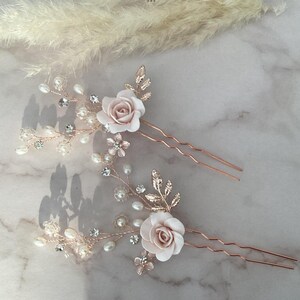 May include: Two rose gold hair pins with delicate floral designs. Each pin features a pink rose, pearl accents, and crystal embellishments.