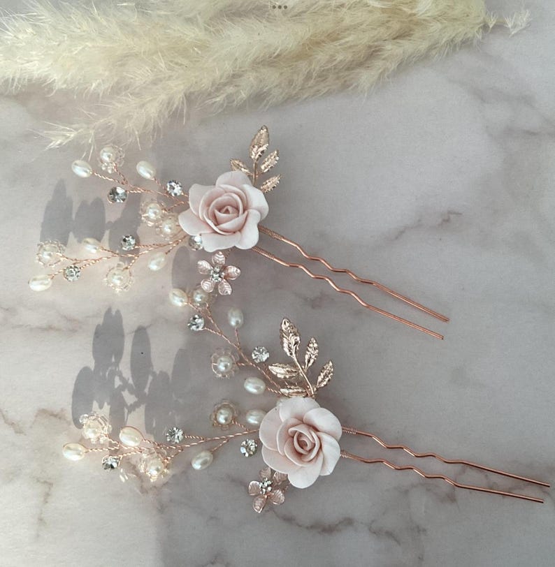 May include: Two rose gold hair pins with delicate floral designs. Each pin features a pink rose, pearl accents, and crystal embellishments.