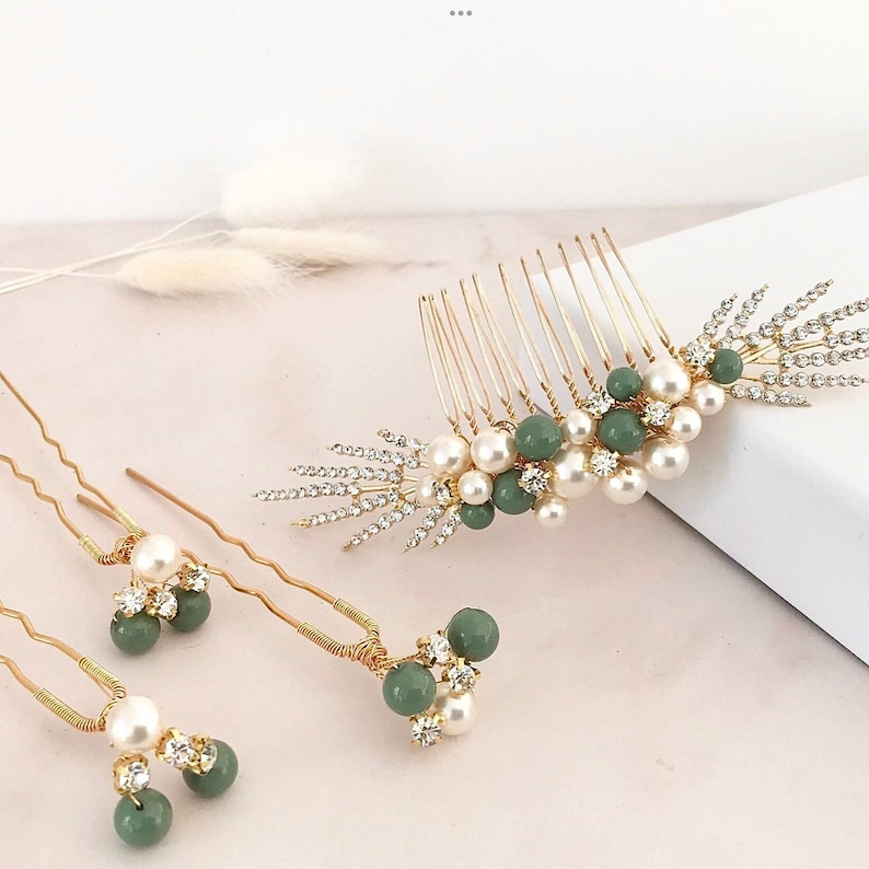 K&ouml;nnte beinhalten: Ein Set aus drei goldenen Haark&auml;mmen mit gr&uuml;nen und wei&szlig;en Perlenakzenten. Der gr&ouml;&szlig;te Kamm hat ein dekoratives Design mit Strasssteinen. Die kleineren K&auml;mme sind Haarnadeln mit einer einzelnen Perle und einer gr&uuml;nen Perle.