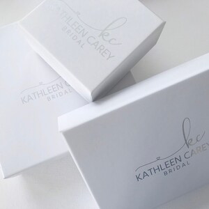 K&ouml;nnte beinhalten: Drei wei&szlig;e Boxen mit dem Logo "kc Kathleen Carey Bridal" in Silber auf der Oberseite jeder Box.