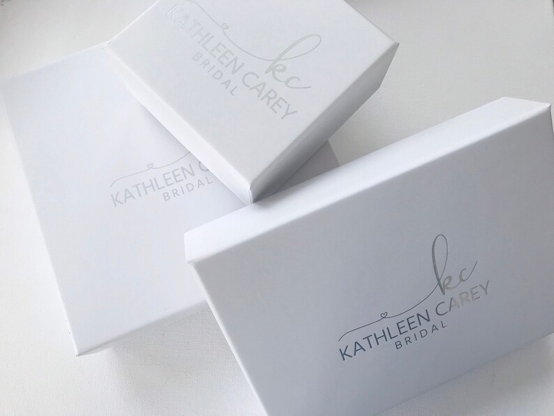 K&ouml;nnte beinhalten: Drei wei&szlig;e Boxen mit dem Logo "kc Kathleen Carey Bridal" in Silber auf der Oberseite jeder Box.