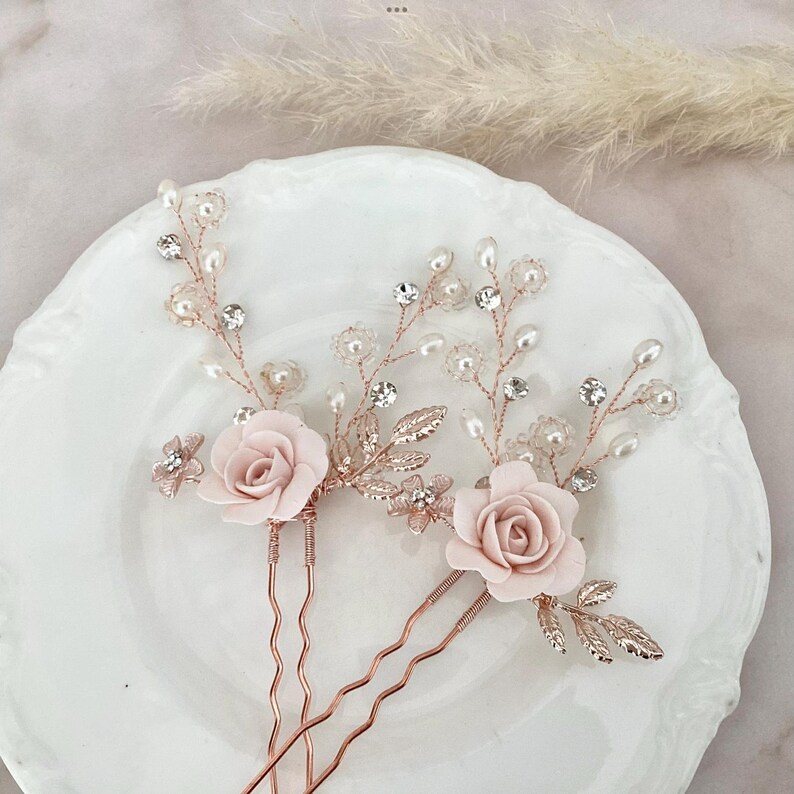 May include: Three rose gold hair pins with pink fabric roses and pearl and crystal embellishments.
